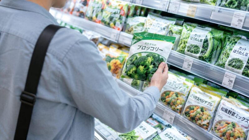 スーパーの冷凍食品売り場で、男性が冷凍ブロッコリーを手に取って選んでいる様子。