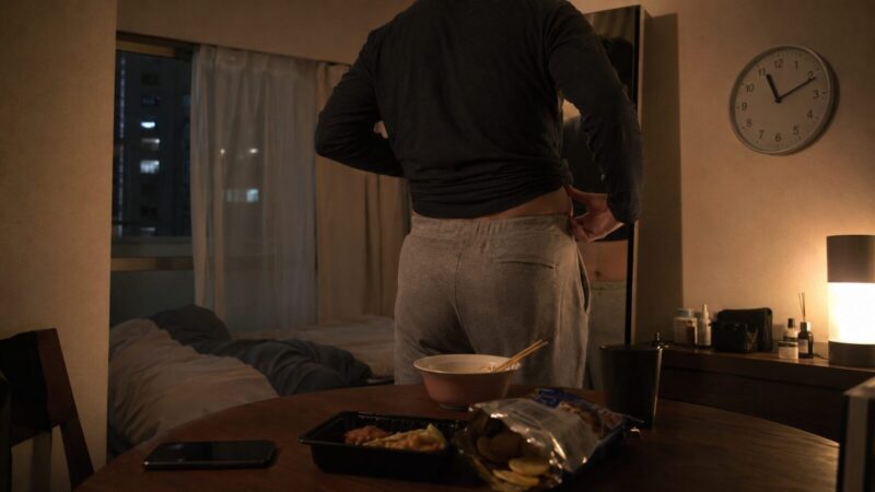 夜のワンルームで、食後に鏡の前に立ち腹回りを気にする男性の後ろ姿。テーブルには夜食やスマートフォンが置かれ、睡眠不足と生活の乱れを感じさせる場面。