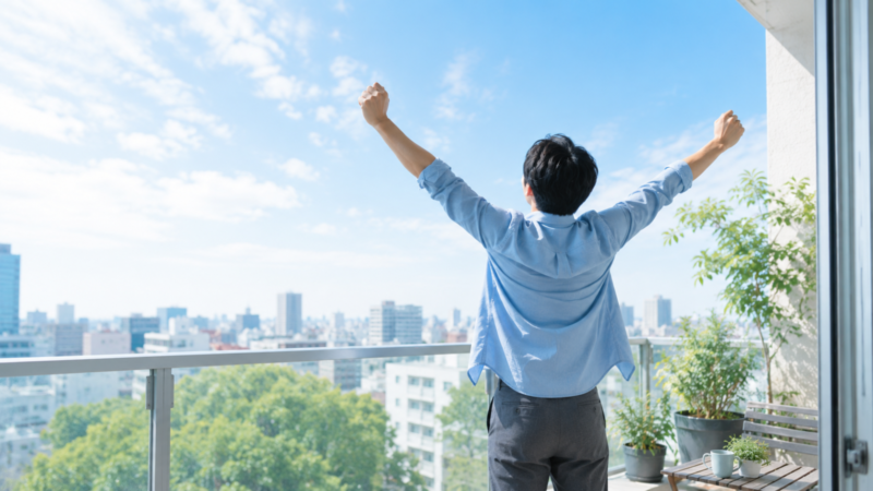 ベランダで大きく伸びをしながら青空を見上げる30代男性の後ろ姿