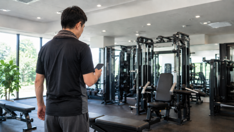 明るいスポーツジムで、スマートフォンを確認しながらトレーニングを始める男性の後ろ姿。